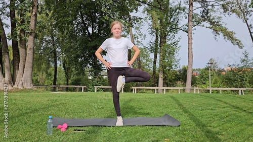 Girl in white t‑shirt and black leggings doing balance exercise on trainer. Pink dumbbells and water bottle on grass. Park with trees, natural daylight
