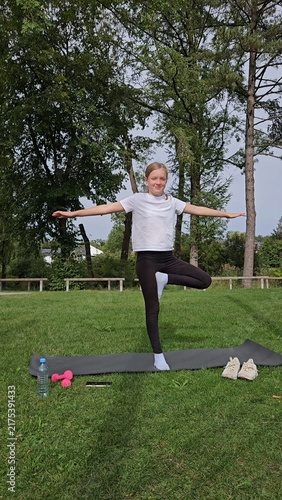 Girl in white t‑shirt and black leggings holding balance pose on trainer. Pink dumbbells and water bottle on grass. Park with trees, bright daylight