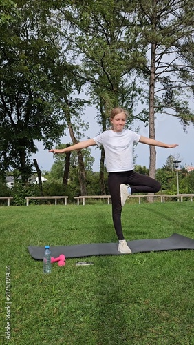 Girl in white t‑shirt and black leggings holding balance pose on trainer. Pink dumbbells and water bottle on grass. Park with trees, bright daylight