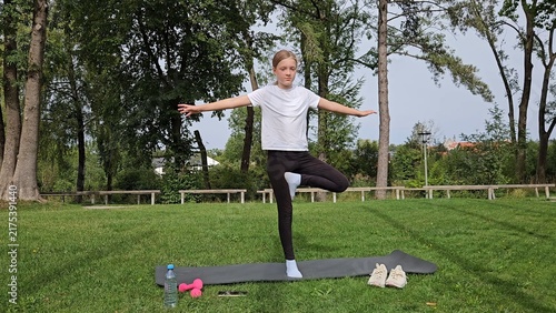 Girl in white t‑shirt and black leggings holding wide balance pose on trainer. Pink dumbbells and water bottle on grass. Park with tall trees, bright daylight
