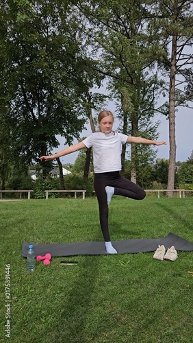 Girl in white t‑shirt and black leggings holding balance pose on trainer. Pink dumbbells and water bottle on grass. Park with trees, bright daylight