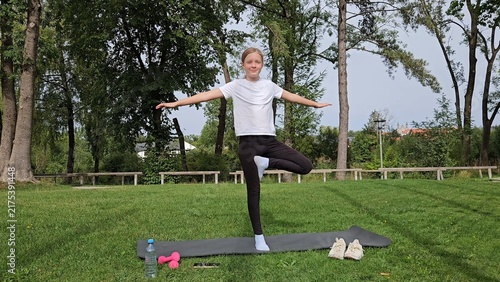 Girl in white t‑shirt and black leggings holding wide balance pose on trainer. Pink dumbbells and water bottle on grass. Park with tall trees, bright daylight