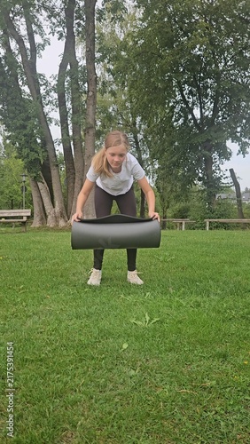 Young girl in a white t‑shirt and dark leggings playfully interacts with a large rolled‑up grey fitness mat in a park. She lifts, holds, and poses with the mat in different frames. The setting feature