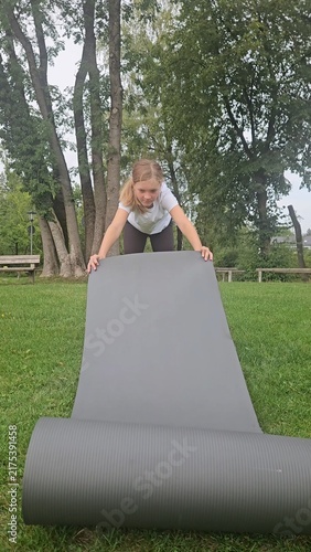Young girl in a white t‑shirt and dark leggings playfully interacts with a large rolled‑up grey fitness mat in a park. She lifts, holds, and poses with the mat in different frames. The setting feature