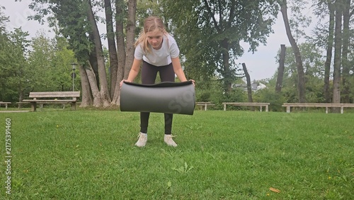 Young girl in a white t‑shirt and dark leggings playfully interacts with a large rolled‑up grey fitness mat in a park. She lifts, holds, and poses with the mat in different frames. The setting feature