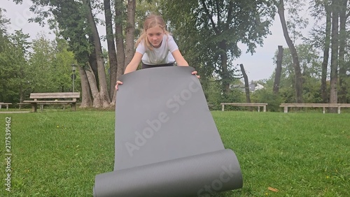 Young girl in a white t‑shirt and dark leggings playfully interacts with a large rolled‑up grey fitness mat in a park. She lifts, holds, and poses with the mat in different frames. The setting feature
