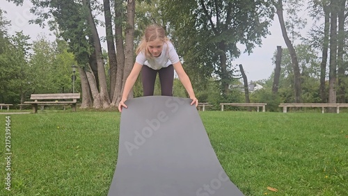 Young girl in a white t‑shirt and dark leggings playfully interacts with a large rolled‑up grey fitness mat in a park. She lifts, holds, and poses with the mat in different frames. The setting feature