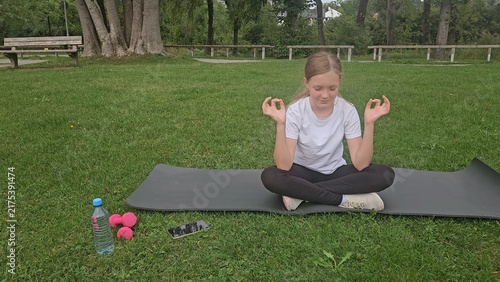 Young girl with light‑blonde hair in a ponytail tied with a blue scrunchie, sitting cross‑legged on a dark grey yoga mat on grass. She’s wearing a plain white t‑shirt, black leggings, and white sneake