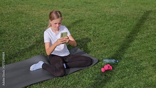 Girl in white t‑shirt and black leggings sitting on grey yoga mat on grass, looking at smartphone. Pink dumbbells and plastic water bottle on grass. Bright daylight, relaxed outdoor atmosphere