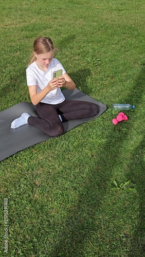 Girl in white t‑shirt and black leggings sitting on grey yoga mat on grass, focused on smartphone. Pink dumbbells and plastic water bottle nearby. Natural daylight, calm outdoor park scene