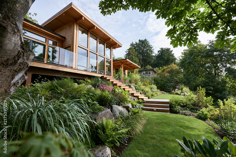 Naklejka premium Modern wooden home blends with lush green surroundings. Stone steps lead to a grassy path lined with native plants. Large windows invite natural light into cozy living spaces. Trees frame the house