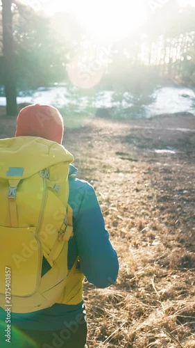 A man walks along a dirt path in a wooded area, carrying a yellow backpack. The man is heading towards sunlight filtering through the trees in a serene landscape.