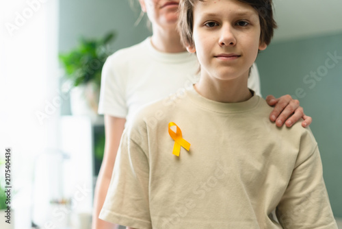 Konstfotografi Vulnerable child wearing yellow ribbon for cancer awareness month is tenderly embraced by loving mother in home interior, showing protective support