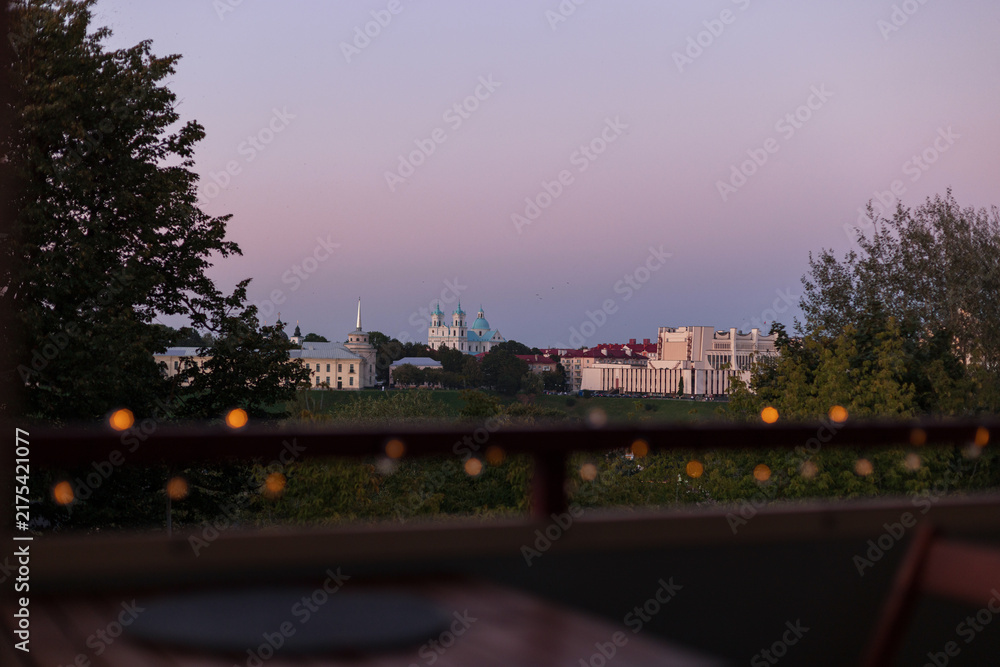 Obraz premium Scenic twilight view over Grodno, Belarus city skyline, featuring St. Francis Xavier Cathedral and Regional Drama Theatre, viewed through a railing with warm bokeh lights at dusk from a terrace.