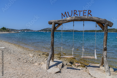 Wooden rope swing with the Murter sign on the pebble shore of a calm bay, Murter island, Croatia