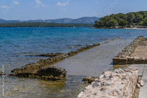 Ancient stone walls of Colentum sinking into the Adriatic shallows below a pine covered shore, Murter, Croatia
