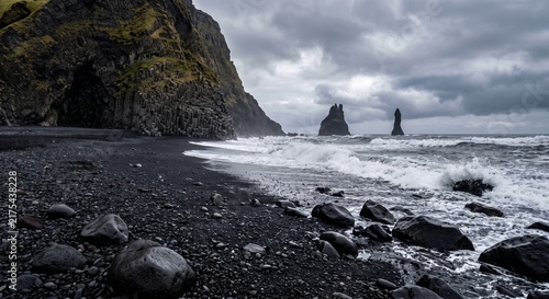Black sand beach with waves and rocky cliffs under a cloudy sky  