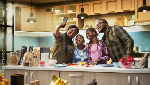 Obraz na plátně Mother takes phone selfies while her family cooks homemade pizza in a modern kitchen, capturing laughter, togetherness and warm family bonding as the children and parents enjoy cooking together