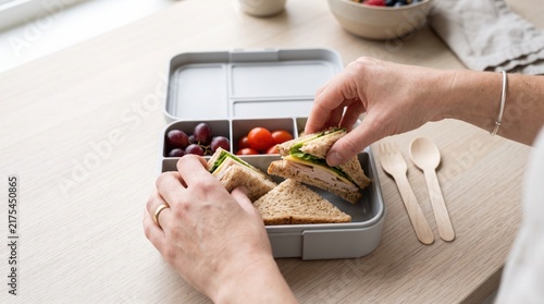 Hands placing a fresh sandwich into a grey lunchbox with fruit and vegetables