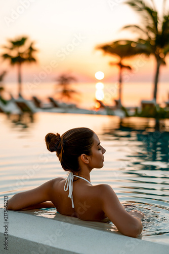 Beautiful young woman relaxing in an infinity pool during a gorgeous tropical sunset with palm trees and ocean view at a luxury summer resort hotel