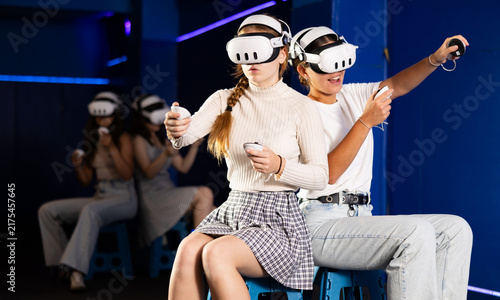 Foto Excited young female friends having fun in modern virtual reality room, playing pair versus pair game in VR glasses, manipulating objects or shooting at targets with gaming controllers in hands