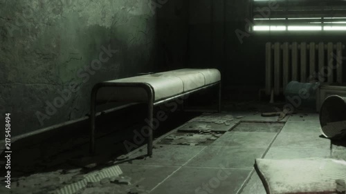 A room shows a metal bench in the center with debris scattered on the floor. Light filters through the slatted window adding shadows on the walls. The atmosphere feels neglected and empty.