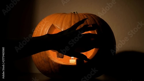 Creepy witch hand reaching towards glowing jack o lantern carved pumpkin in dark room for horror halloween holiday atmosphere