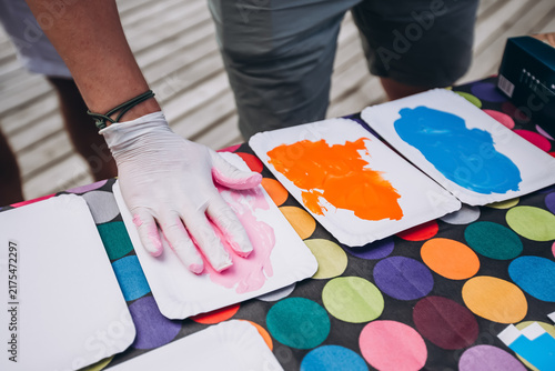 Quadro su tela Finger Painting Fun: A gloved hand dips into vibrant pink paint, ready to create art on a paper plate, surrounded by a colorful array of other paints and a playful polka dot tablecloth