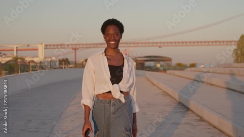 Black African American woman walks along a waterfront promenade during golden hour, smiling while enjoying an evening outdoors.