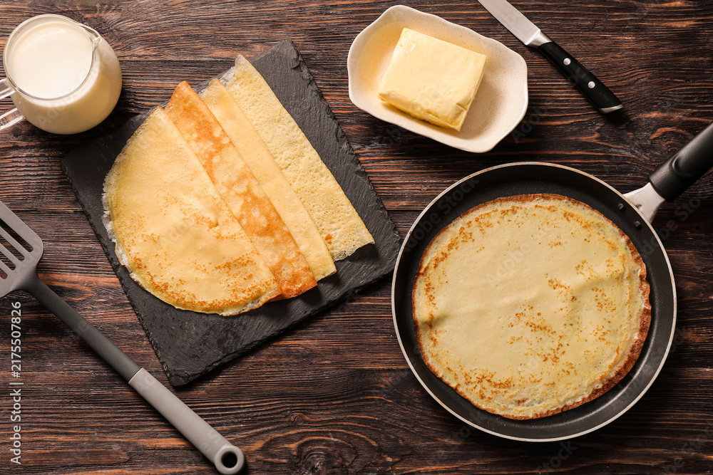Obraz premium Frying pan with tasty thin pancakes, spatula, knife and ingredients on wooden background