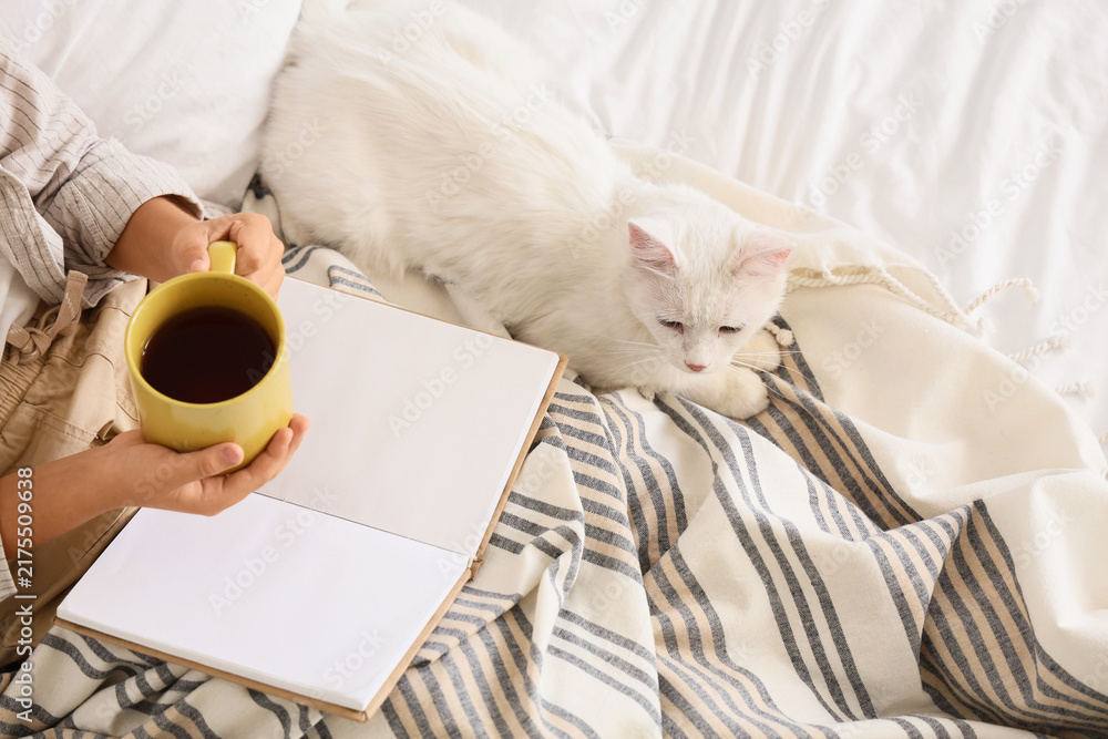Naklejka premium Little boy with cup of tea and cute cat reading book under blanket on bed, top view