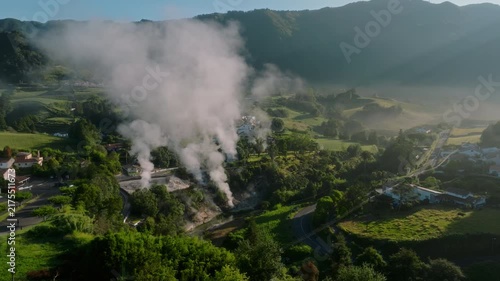 Aerial perspective of the Furnas valley on Sao Miguel Island, showcasing billowing geothermal steam rising against a backdrop of sun-drenched green hills and a peaceful rural village.