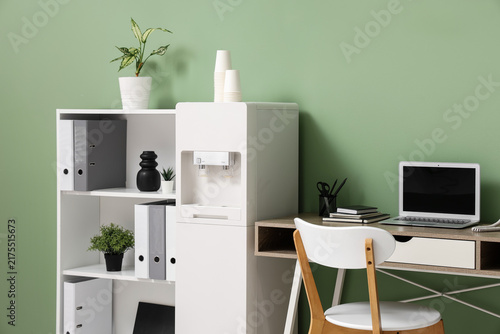 Interior of stylish office with modern water cooler, workspace and shelving unit