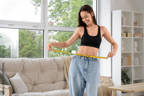 Beautiful young happy woman with measuring tape wearing loose jeans in living...
