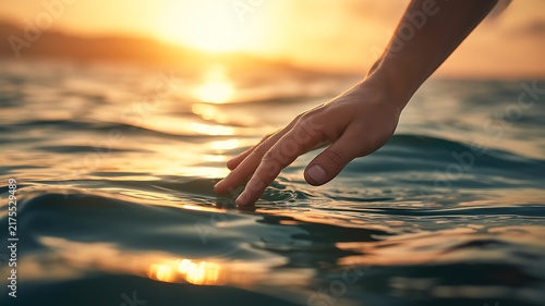Human hand reaching down to touch the surface of calm ocean water at golden hour sunset, ripples