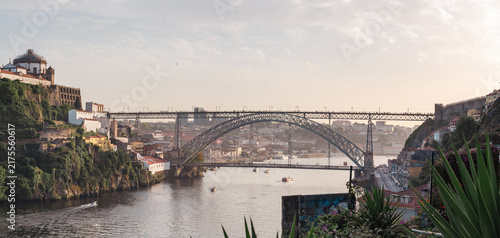 Scenic panoramic view of the Dom Luís I Bridge spanning the Douro River in Porto, Portugal, with historic riverside architecture, lush hillsides, boats, and warm golden-hour light.