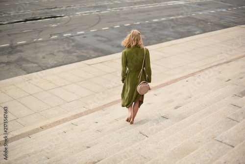 Rear view of stylish blonde woman in green coat walking down wide stone stairs in city