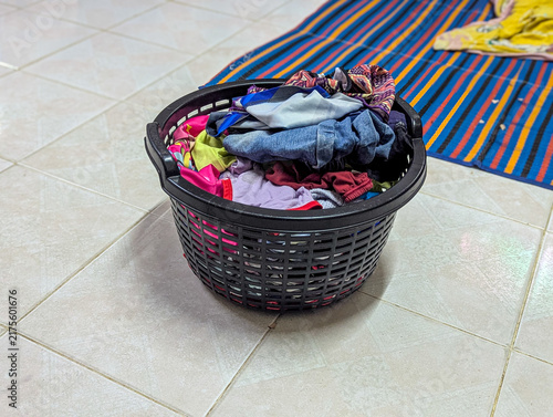 Black plastic laundry basket filled with clothes on tiled floor.
