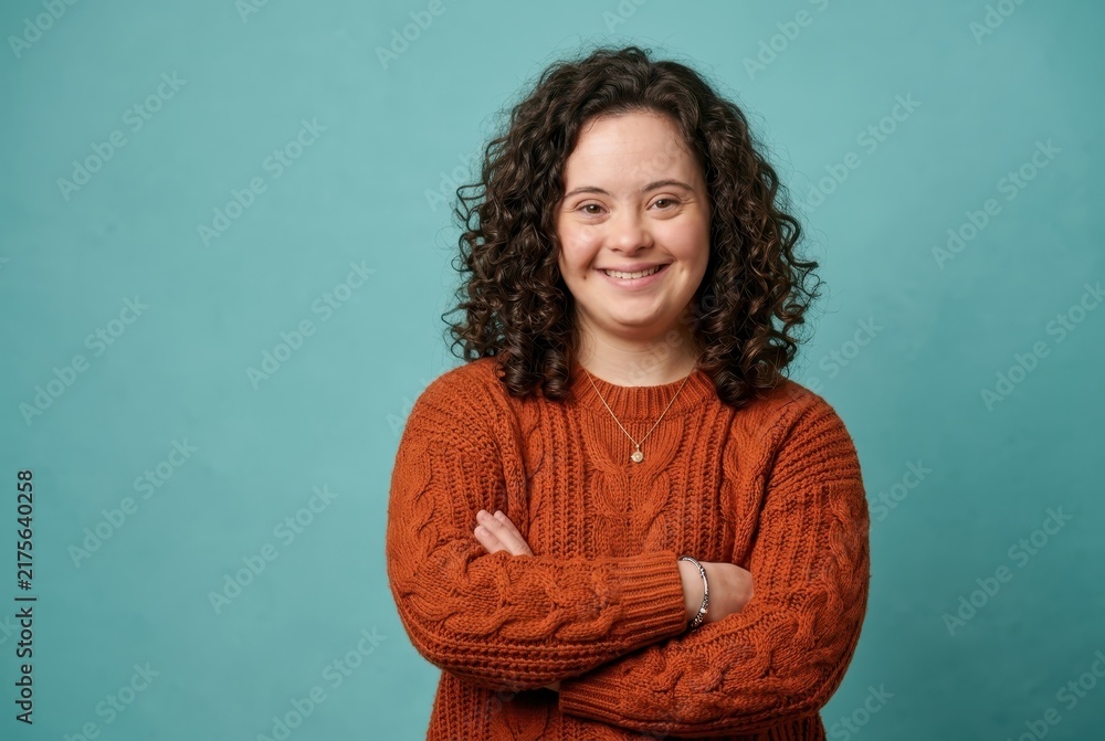 Obraz premium Confident Young Woman with Down Syndrome Smiling with Arms Crossed Against Teal Background