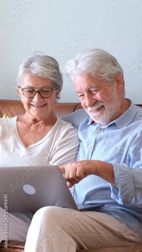 Happy mature caucasian couple sitting on the sofa at home, laughing and having fun while using a laptop computer, enjoying retirement and technology together in their living room