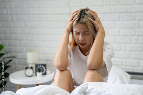 Stressed Asian woman sits on the bed, placing her free hand on her head unhappy female spending early morning in bedroom at home