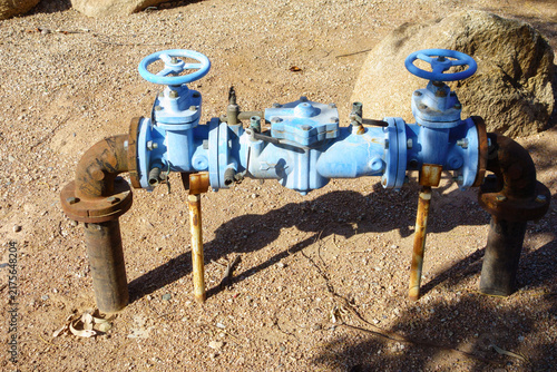 Blue painted water backflow preventer assembly with two handwheel shutoff valves and rusty plumbing pipes on ground outdoors