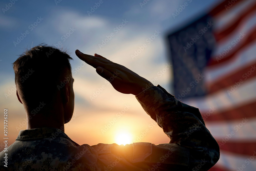 Fototapeta premium Soldier salutes at sunset, flag blurred in background. Backlit figure shows solemn commitment and pride. Golden light glows behind disciplined posture. Ideal for military, patriotism