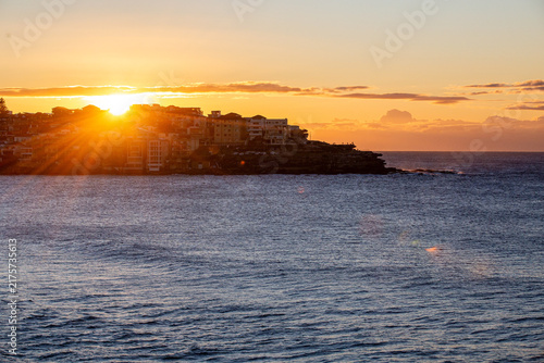 Golden sunset over ocean beside a densely developed coastal headland with dramatic sun rays