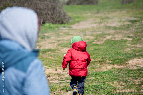 A child is running in a green meadow. The boy is spending time in nature with his family. A boy in a red jacket is having fun in nature. People, model, person. 