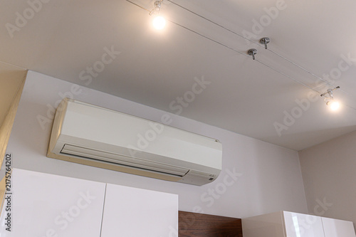 Wall‑mounted white split‑system air conditioner above glossy white cabinets. minimalist room with track lighting, clean lines, dark wood accent panel