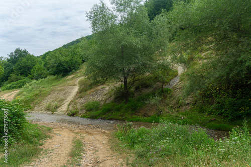 A dirt road leading to a shallow river. A steep bank overgrown with trees and bushes. Hills and a cloudy sky in the background.