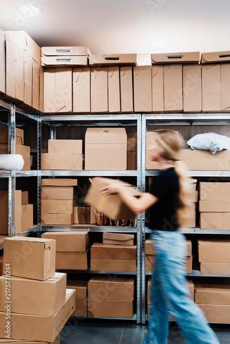 Obraz na płótnie Blurred woman in motion pulls a cardboard box from metal shelving in a small storage room packed with parcels