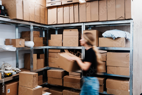 Fotografie Blurred worker carries a cardboard box past metal shelves stacked with parcels in a small storage room