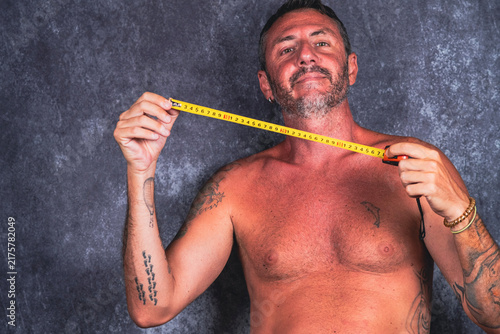 A man holds a tape measure. The man is bare-chested, has short hair and a beard